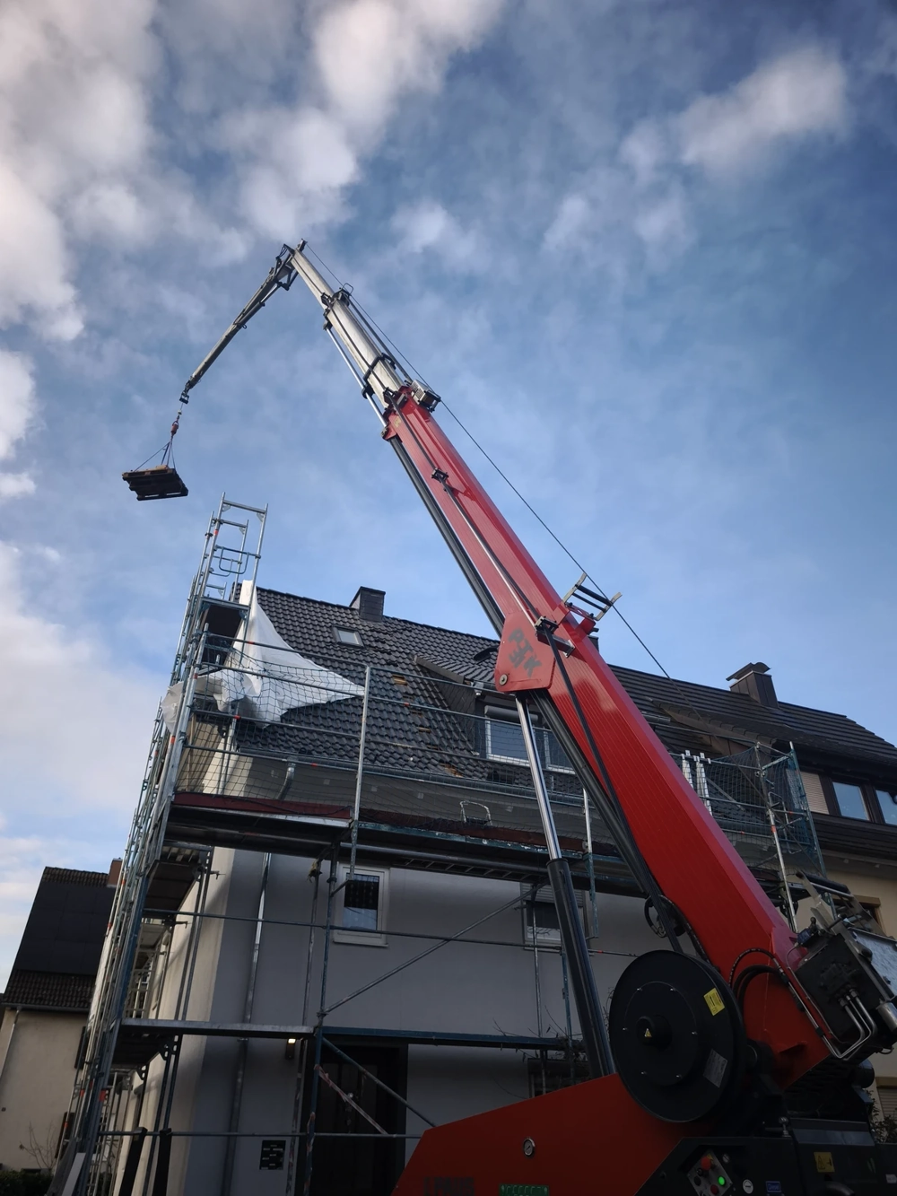 Mobilkran hebt Paletten mit Dachmaterialien an ein eingerüstetes Wohnhaus zur Dachsanierung