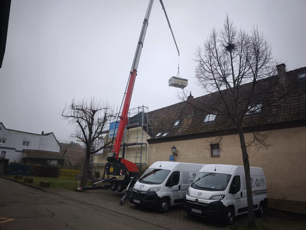Roter Autokran hebt Dachbaustoffe auf Haus mit Gerüst und Firmenwagen bei Dachsanierung