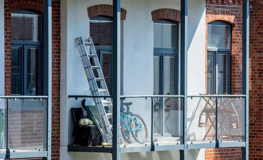 Metallleiter und Fahrrad auf Glas-Geländer-Balkon vor roter Backsteinfassade – Balkonsanierung