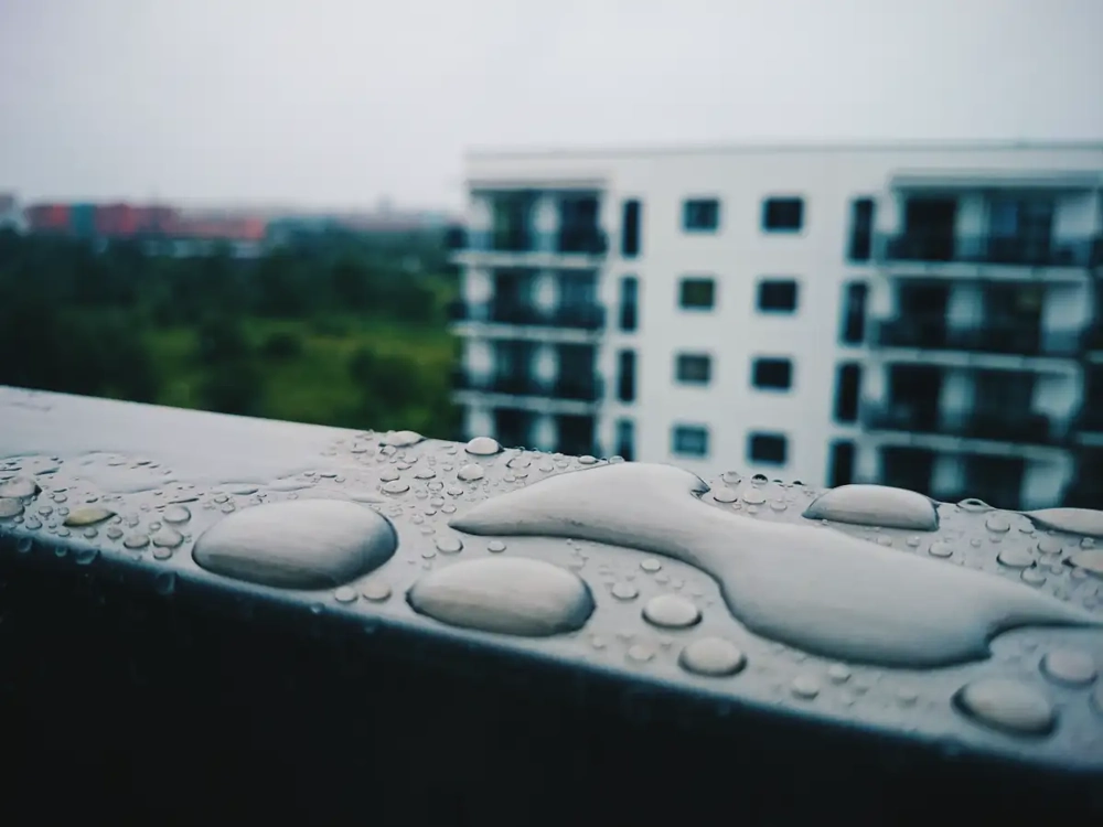 Nahaufnahme von Wassertropfen auf einem Metallgeländer, verschwommener Wohnkomplex im Hintergrund